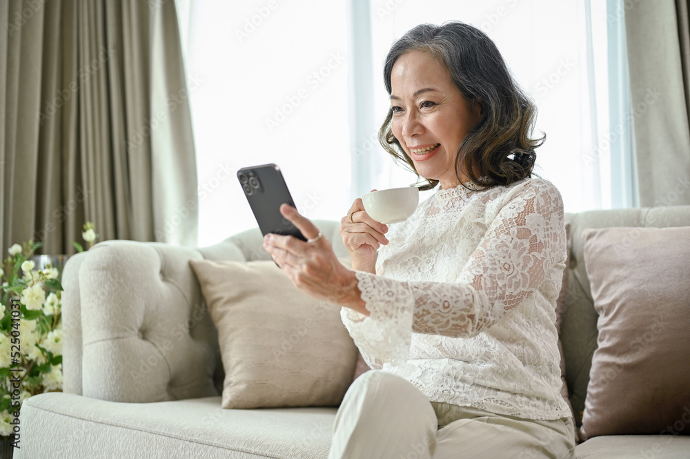 Gorgeous Asian-aged woman relaxes in the living room, sipping coffee, using smartphone Gorgeous Asian-aged woman relaxes in the living room, sipping coffee, using smartphone