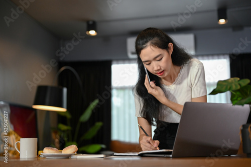 Attractive young Asian female taking notes on her notepad while talking on the phone