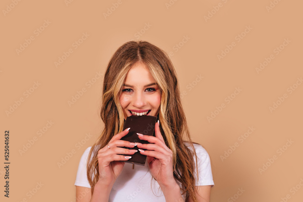 Smiling cheerful teenage girl with long curly hair in a white t-shirt eating a bar of chocolate isolated on a beige background. Copy space. Smiling cheerful teenage girl with long curly hair in a white t-shirt eating a bar of chocolate isolated on a beige background. Copy space.
