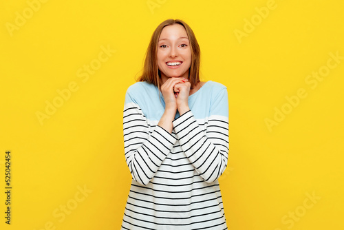 Cute fair-haired female, holding hands under chin, has amazed and happy expression, being excited, expresses positive emotions, standing in striped sweatshirt over yellow background