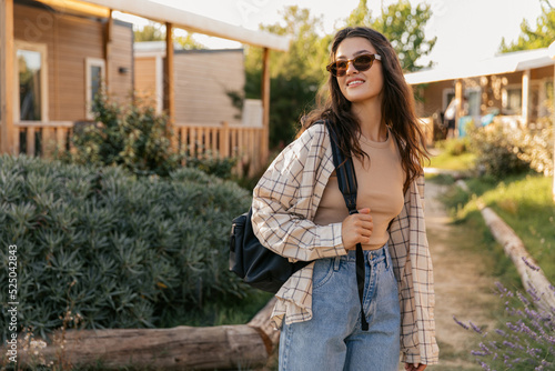 Fashionable caucasian young girl posing relaxing outdoors outside city. Brunette wears sunglasses, tank top, shirt, jeans and backpack. Concept of enjoying moment