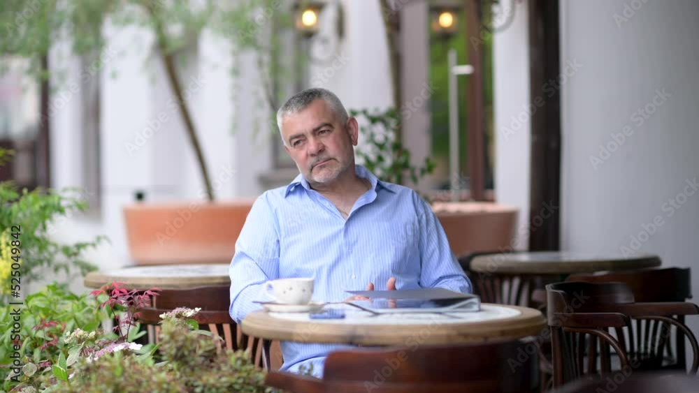 Handsome caucasian middle aged man sitting at the street cafe with a cup of coffee or tea on the table. Man sitting at table in street cafe looks around enjoying morning coffee. People lifestyle conce Handsome caucasian middle aged man sitting at the street cafe with a cup of coffee or tea on the table. Man sitting at table in street cafe looks around enjoying morning coffee. People lifestyle conce