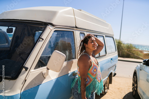 Chica joven guapa posando y sonriendo delante de furgoneta vintage en aparcamiento de la playa photo
