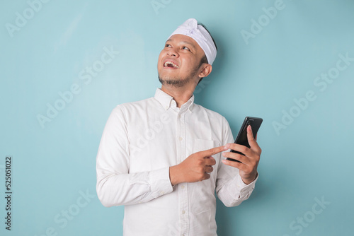 Portrait of an excited young Balinese man wearing udeng or traditional headband and white shirt looking aside while holding smartphone isolated over blue background