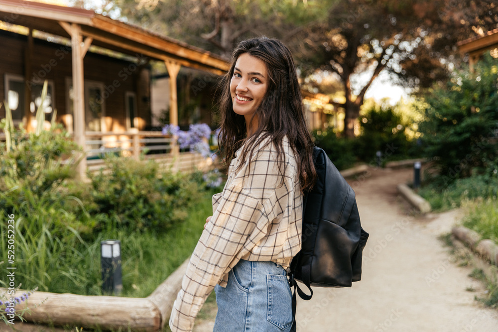 Pretty young caucasian woman walks in countryside at weekend alone. Brunette smiling looking at camera, wears shirt, jeans and backpack. Happy weekend concept.