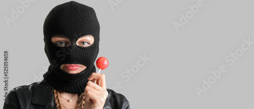 Fotografia Young woman in balaclava and with lollipop on grey background with space for tex
