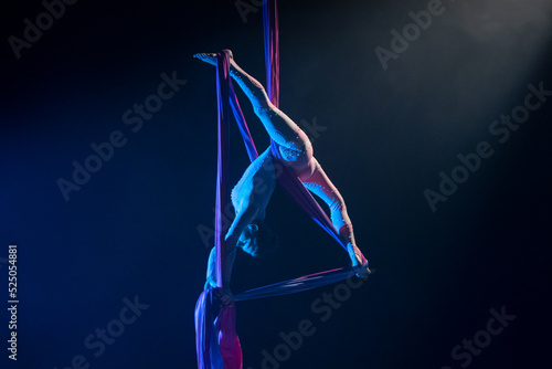 Female circus gymnast hanging upside down on aerial silk and demonstrates stretching. Young woman performs tricks at height on silk fabric. Acrobatic stunts on black background with blue backlight.