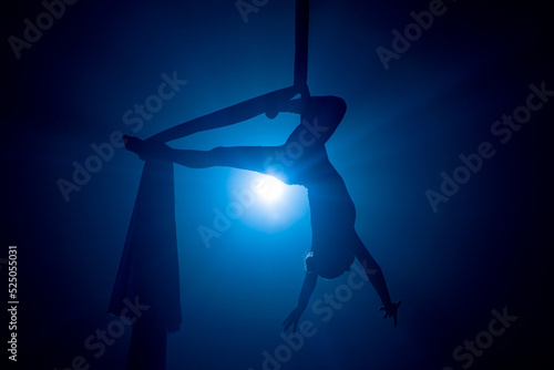 A young woman performing in a circus on aerial silk in the dark with blue light. Silhouette of a female equilibrist hanging upside down and demonstrating the twine. Performance of aerial gymnast.