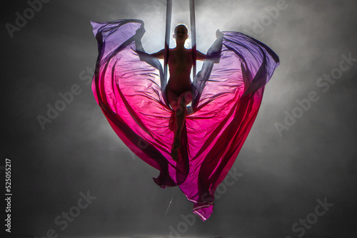 Silhouette of a young woman balancing on an airy silk ribbon and waving a cloth like butterfly wings. A female athlete performing in a circus at a height in the dark with backlight.