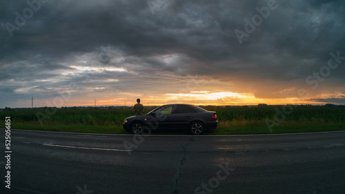 The concept of traveling by car. A young man is resting from the road, sitting on the hood of his car. Sunset over the field in cloudy rainy weather