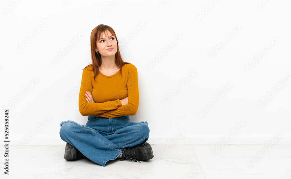 Redhead girl sitting on the floor isolated on white background looking up while smiling