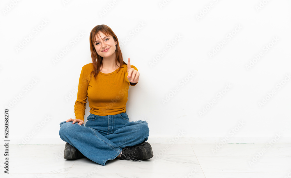 Redhead girl sitting on the floor isolated on white background showing and lifting a finger