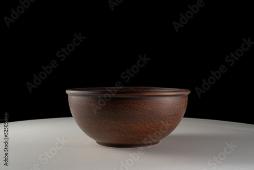 Empty earthen deep bowl on white table isolated on black studio background. Brown handicraft ramekin or rustic plate. Empty earthenware, crockery from clay for food. Old dishes round shape.