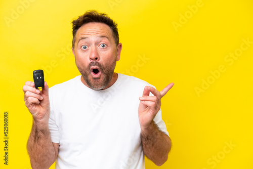 Middle age caucasian man holding car keys isolated on yellow background surprised and pointing side