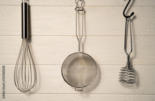 Whisk  masher and strainer hanging against a rustic white wall in restaurant or home kitchen. Kitchen utensils made of stainless steel or metal. Set of tools for cooking food. Kitchenware on drainer.