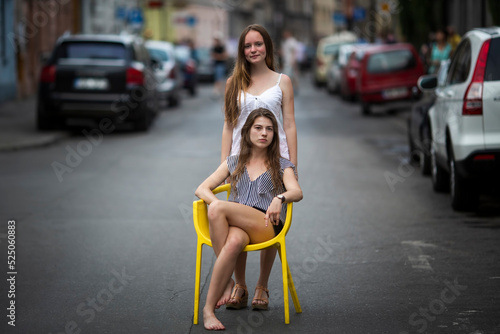 Two pretty girlfriends pose sitting on a chair on an old town street.