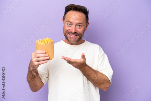 Middle age caucasian man holding fried chips isolated on purple bakcground extending hands to the side for inviting to come © luismolinero