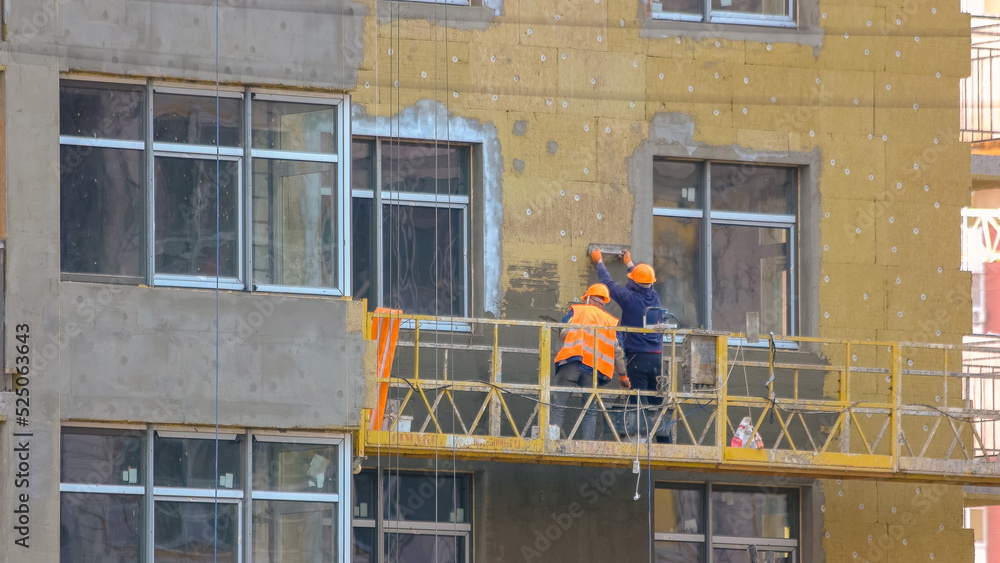 Workers are insulating the wall of a building. Construction site with working men on the elevator. panels. Hanging suspended cradle elevator.