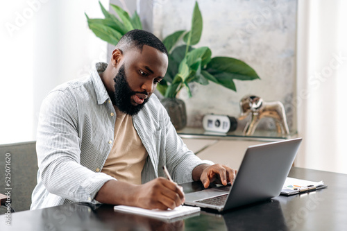 Focused successful smart african american male company owner, manager, sitting at a desk in modern office, working on a project, with documents, analyzing, taking notes, prepares a report