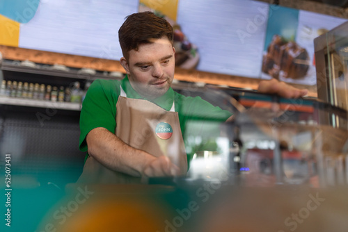 Happy waiter with Down syndrome standing by counter and prepairing hot-dog to a customer in cafe at gas station.