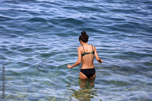 Girl in black swimsuit going to swim in sea water. Vacation on a pebble beach of summer resort