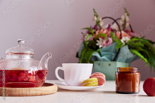 Autumn cozy tea time - cup of tea, glass teapot, candle and macaroons. Herbal raspberry fruit natural tea. Aesthetic home with tea and dessert. Self care, wellness lifestyle.