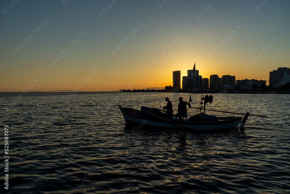 View of fishing boat with two fisherman gliding in the sea at sunset in Mavisehir, Karsiyaka, İzmir. Beautiful sunlight over the city. Selective focus, noise effect and grainy texture. View of fishing boat with two fisherman gliding in the sea at sunset in Mavisehir, Karsiyaka, İzmir. Beautiful sunlight over the city. Selective focus, noise effect and grainy texture.