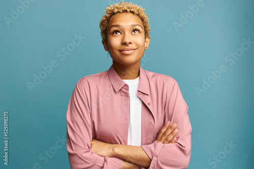 Beautiful romantic female with blond short curly hair in pink silk shirt standing with inspired face expression looking up with dreamy look and crossed hands, recollecting best memories