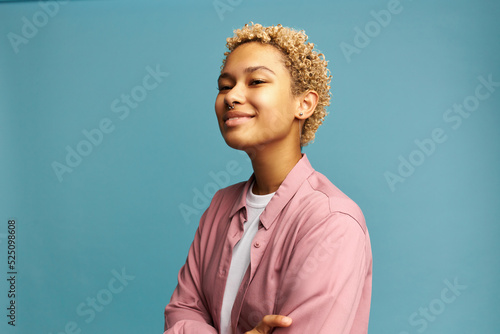 Portrait of narcissistic young blond woman with dark skin and nose piercing standing on blue studio background with lifted chin, feeling proud with herself, having big ego and ambitions