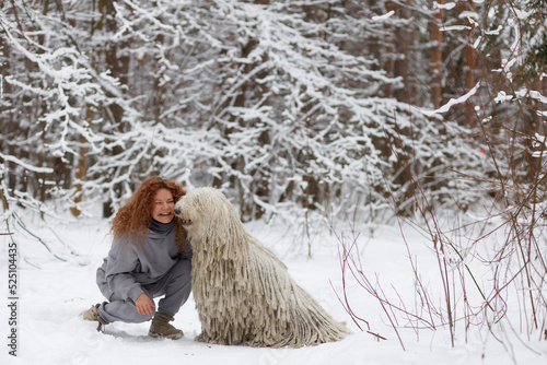 a curly-haired girl sits next to a komondor dog in a winter forest photo