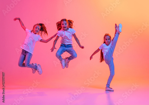 Group of school age girls, cheerful kids in casual style clothes dancing, jumping isolated on pink background in yellow neon light. Music, dance, art