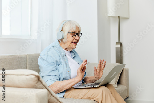a happy, smiling elderly woman is sitting at home on a cozy sofa with headphones on her head, holding a laptop on her lap while working from home © Tatiana