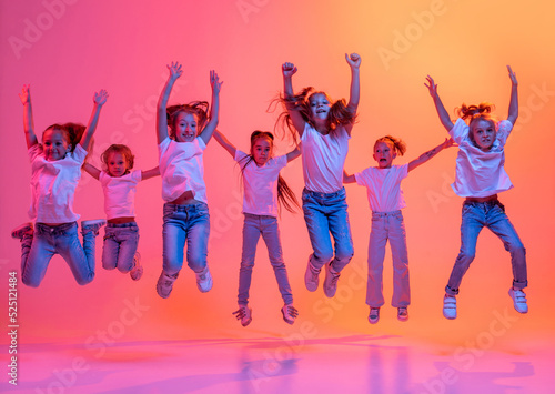 Happy kids, school age girls in white t-shirts and jeans jumping isolated on pink background in yellow neon light.
