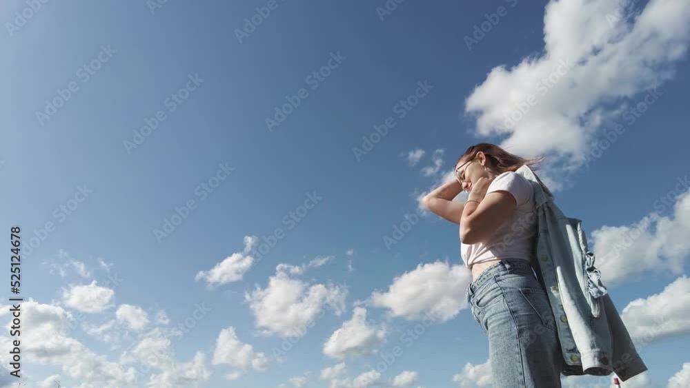 Side view of young woman wearing glasses and jeans standing against blue sky on sunny day enjoying the weather. Carefree woman fixing her red hair hairstyle outdoors. Side view of young woman wearing glasses and jeans standing against blue sky on sunny day enjoying the weather. Carefree woman fixing her red hair hairstyle outdoors.