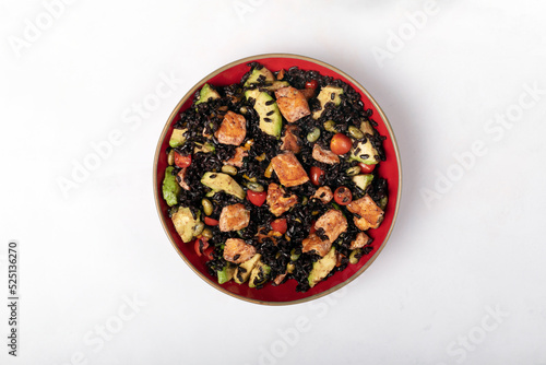 A red bowl of black rice with baked salmon, avocado, tomatoes, chili peppers and edamame.