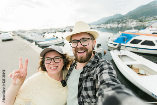 Young couple taking a self portrait laughing as they pose close together for camera on their smartphone outdoors in summer port marina with boats and yachts