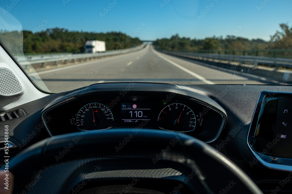 Driver view to the speedometer at110 kmh or 110 mph and the road blurred in motion, day light view from inside a car of driver POV of the road landscape.