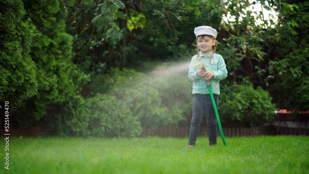A small child is watering the plants near the house with a hose. A cute little girl in a stylish outfit. Garden irrigation. Happy childhood. Daughter helps parents. A small child is watering the plants near the house with a hose. A cute little girl in a stylish outfit. Garden irrigation. Happy childhood. Daughter helps parents.
