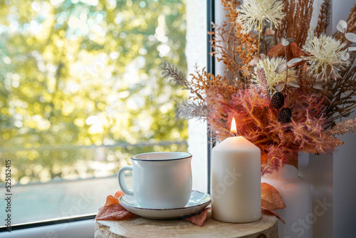 Cup of autumn tea (coffee, chocolate) and yellow dry leaves in the vase, candle near a window, copy space. Hot drink for autumn cold rainy days. Hygge concept, autumn mood