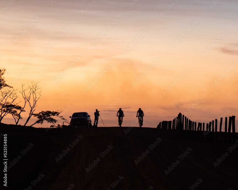Fototapeta premium Silhouette of three cyclists on a rural road with a fence at sunset, orange-yellow sky
