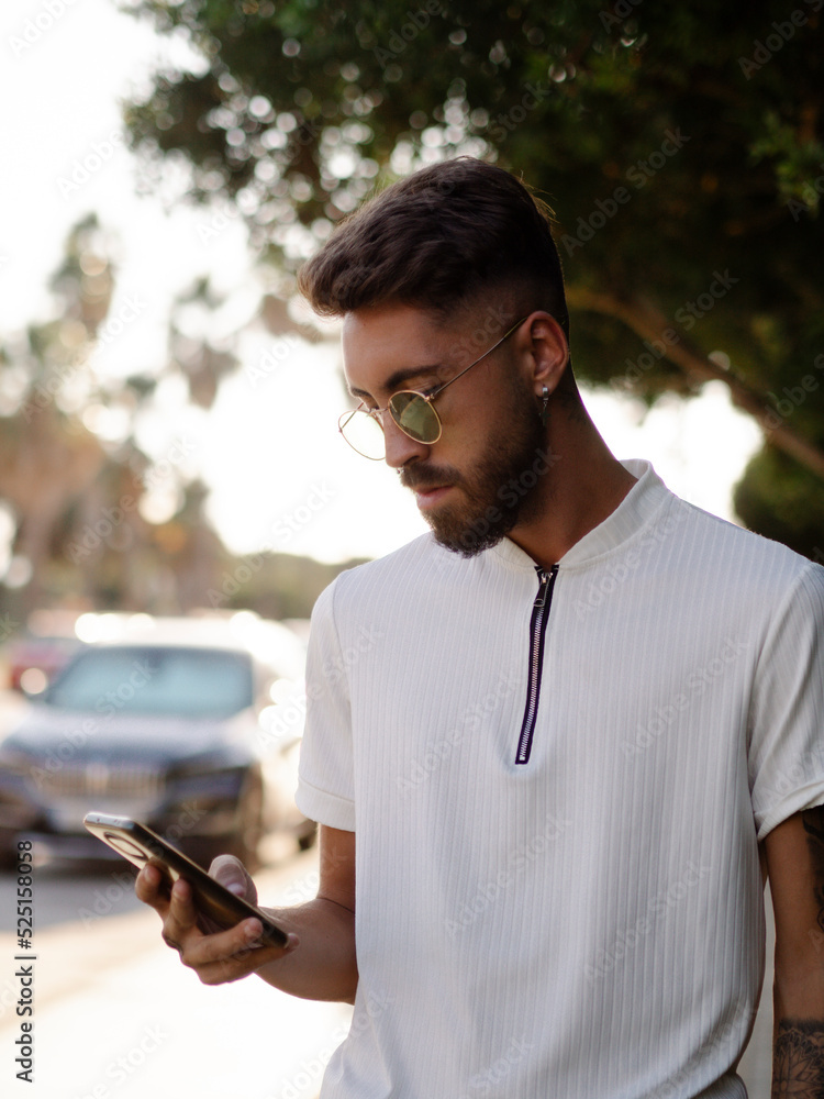 Hombre joven vistiendo gafas de sol usando su teléfono móvil en la calle durante sus vacaciones de verano Hombre joven vistiendo gafas de sol usando su teléfono móvil en la calle durante sus vacaciones de verano