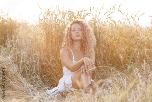 curly-haired beautiful girl dressed in a white sundress in the field basking in the sunset sun