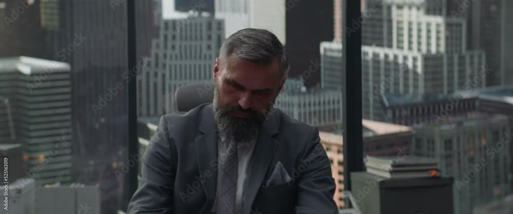 Portrait of 50s adult grey-haired Caucasian businessman signing documents in his office located in a skyscraper, business district buildings in the background Portrait of 50s adult grey-haired Caucasian businessman signing documents in his office located in a skyscraper, business district buildings in the background