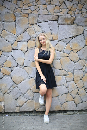 Blonde young woman against stone wall background, smiling and posing on background of vineyards