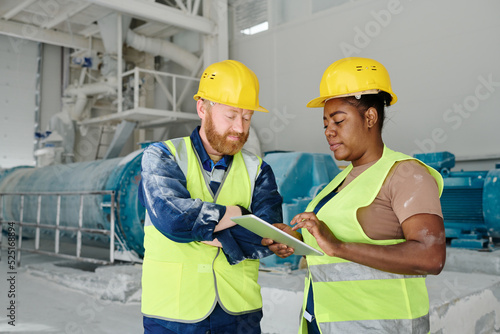 Two intercultural engineers in workwear and safety helmets networking or looking through electronic sketch or construction plan