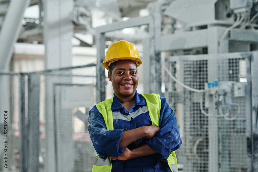 Young successful African American female worker of factory in uniform and protective helmet looking at you while standing in workshop