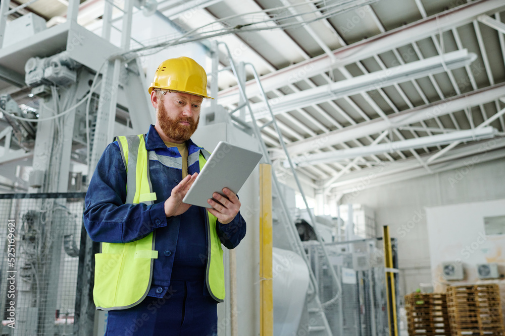 Young male engineer with tablet concentrating on reading online information while standing in spacious workshop of factory Young male engineer with tablet concentrating on reading online information while standing in spacious workshop of factory