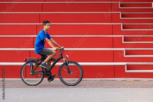 A young teen boy in a blue t-shirt rides a bicycle past a red wall