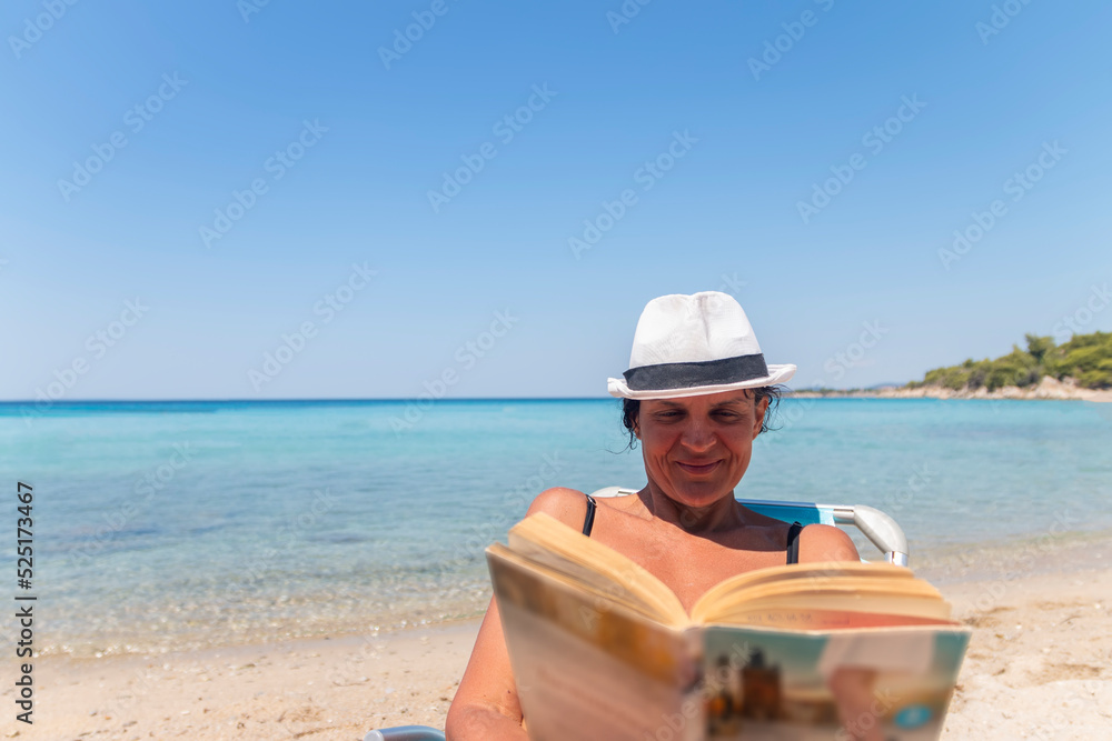 Portrait of a happy woman sitting on a deckchair and reading a book. Happy woman on the beach with a beautiful view. Summer vacation concept.
