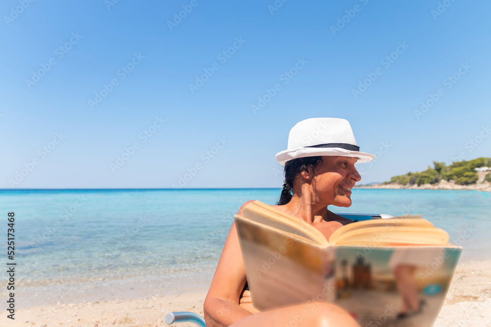 Portrait of a happy woman sitting on a deckchair and reading a book. Happy woman on the beach with a beautiful view. Summer vacation concept.